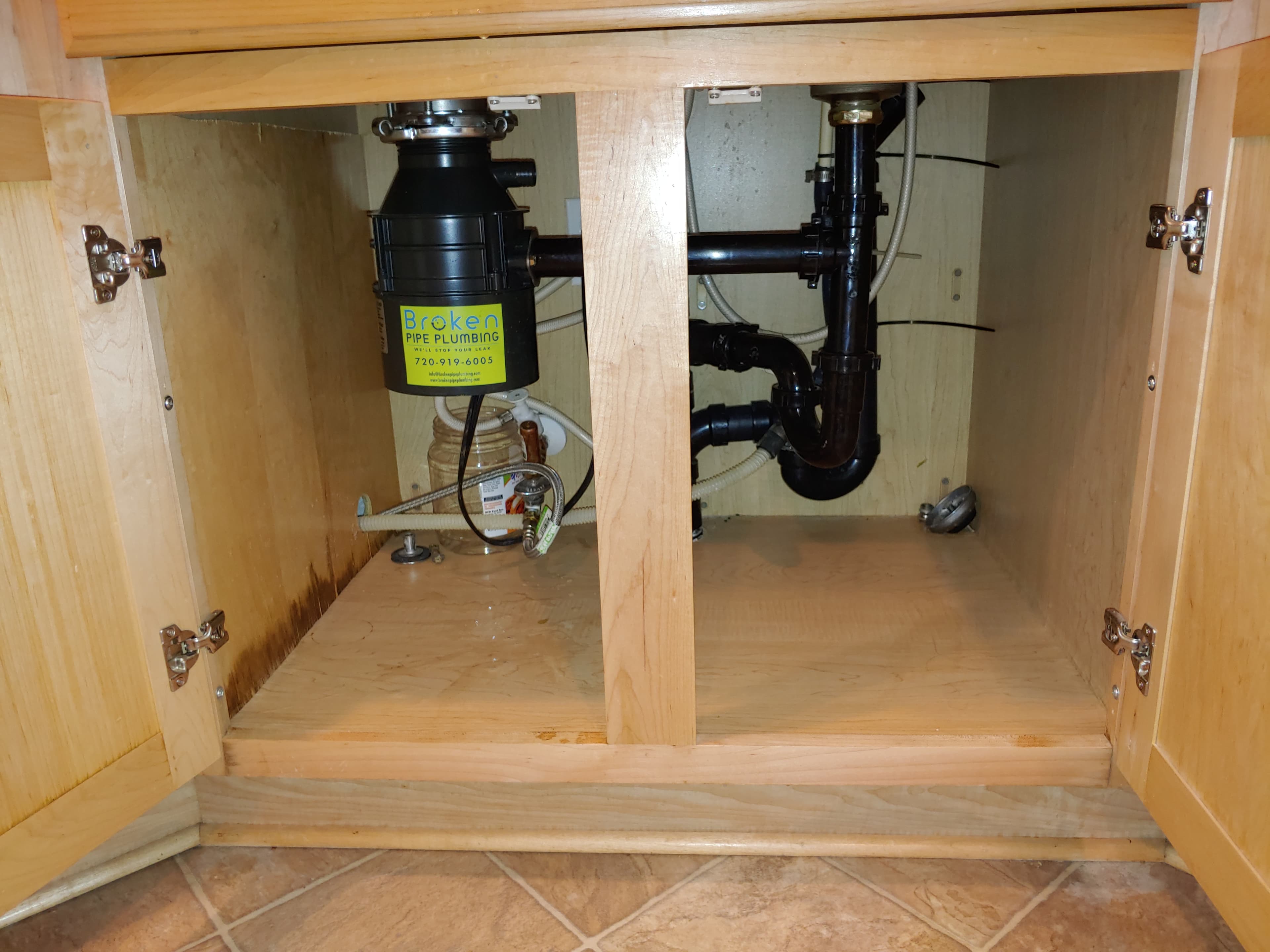 Kitchen sink cabinet with a newly installed garbage disposal and black drain lines, labeled with a Broken Pipe Plumbing sticker