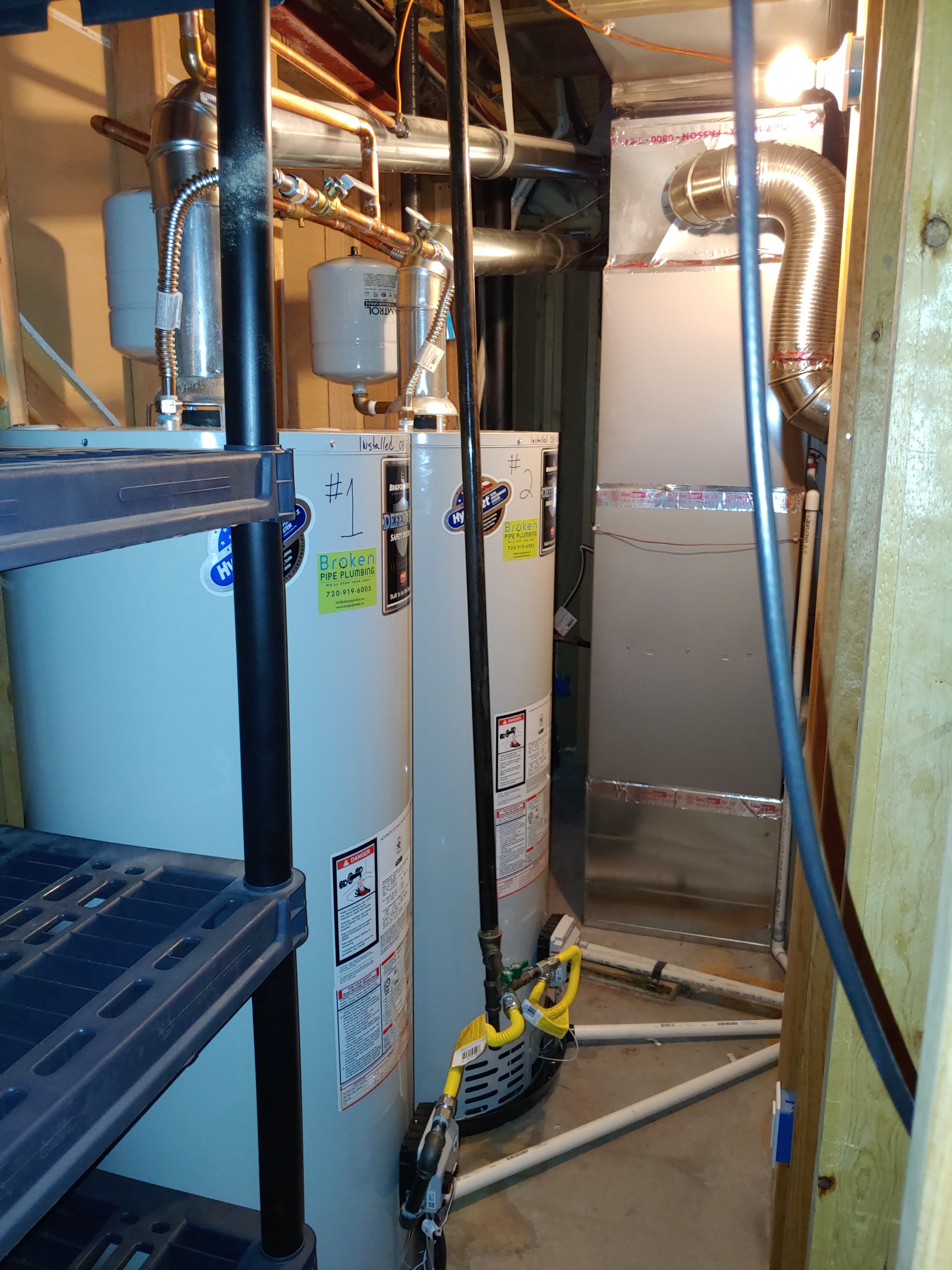 Two water heaters installed side by side in a basement, each with a Broken Pipe Plumbing sticker